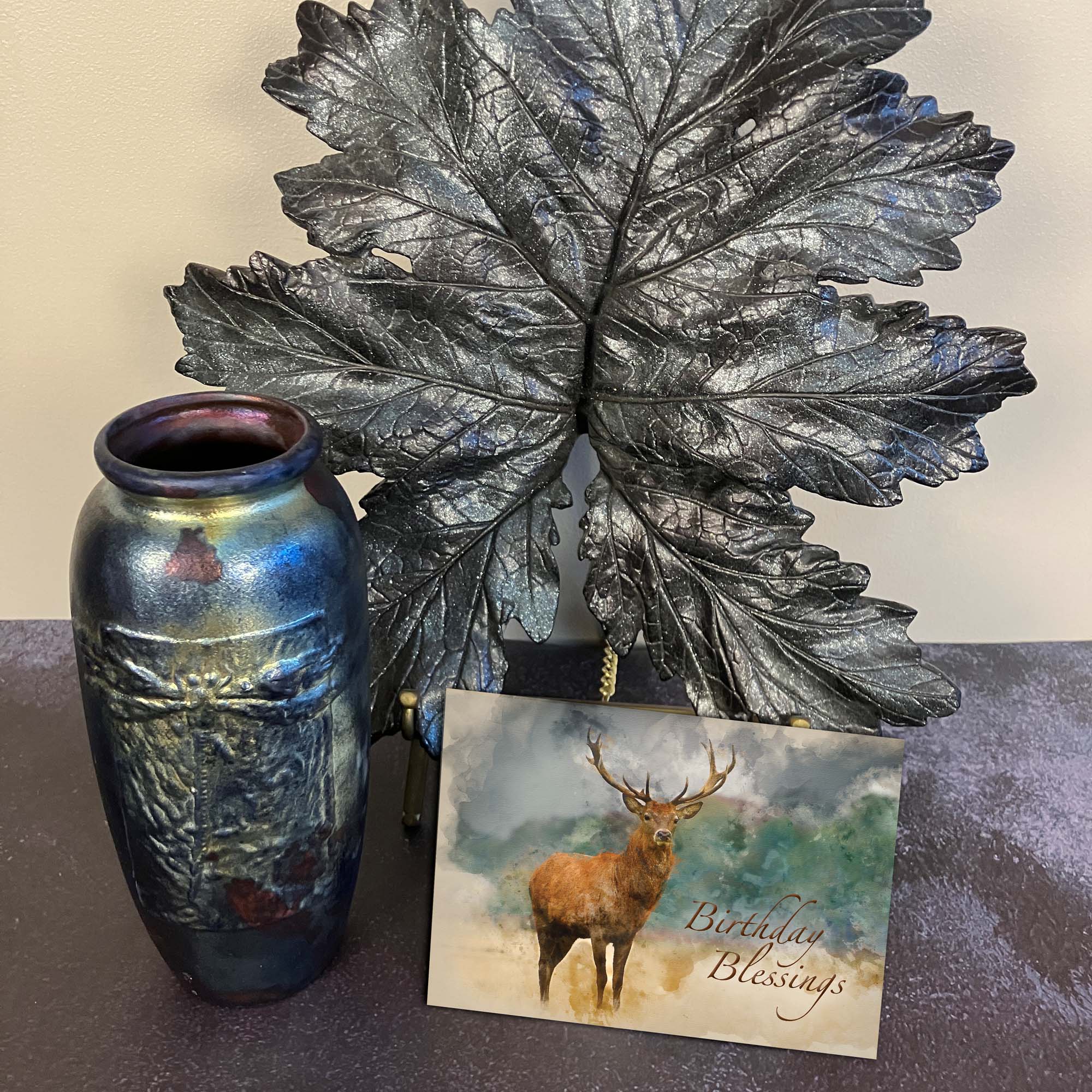 Decorative vase, metallic leaf wall art, and card with deer illustration on a dark surface.