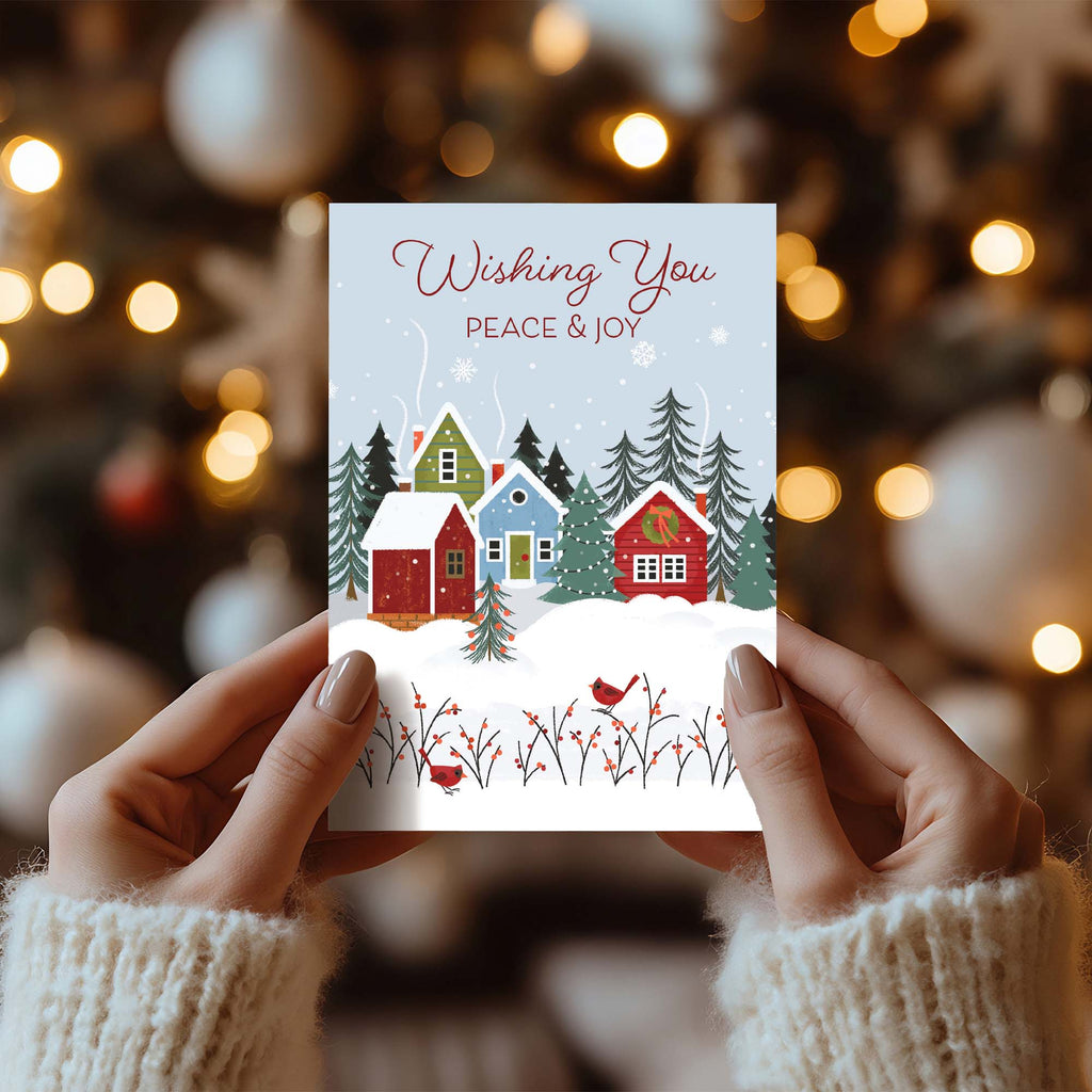 Christmas card with snowy village scene held by hands against a blurred festive background