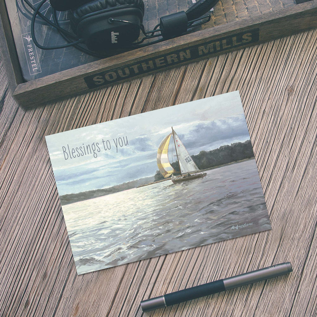 Greeting card with sailboat on water and 'Blessings to you' text, placed on a wooden surface with headphones and a box in the background.