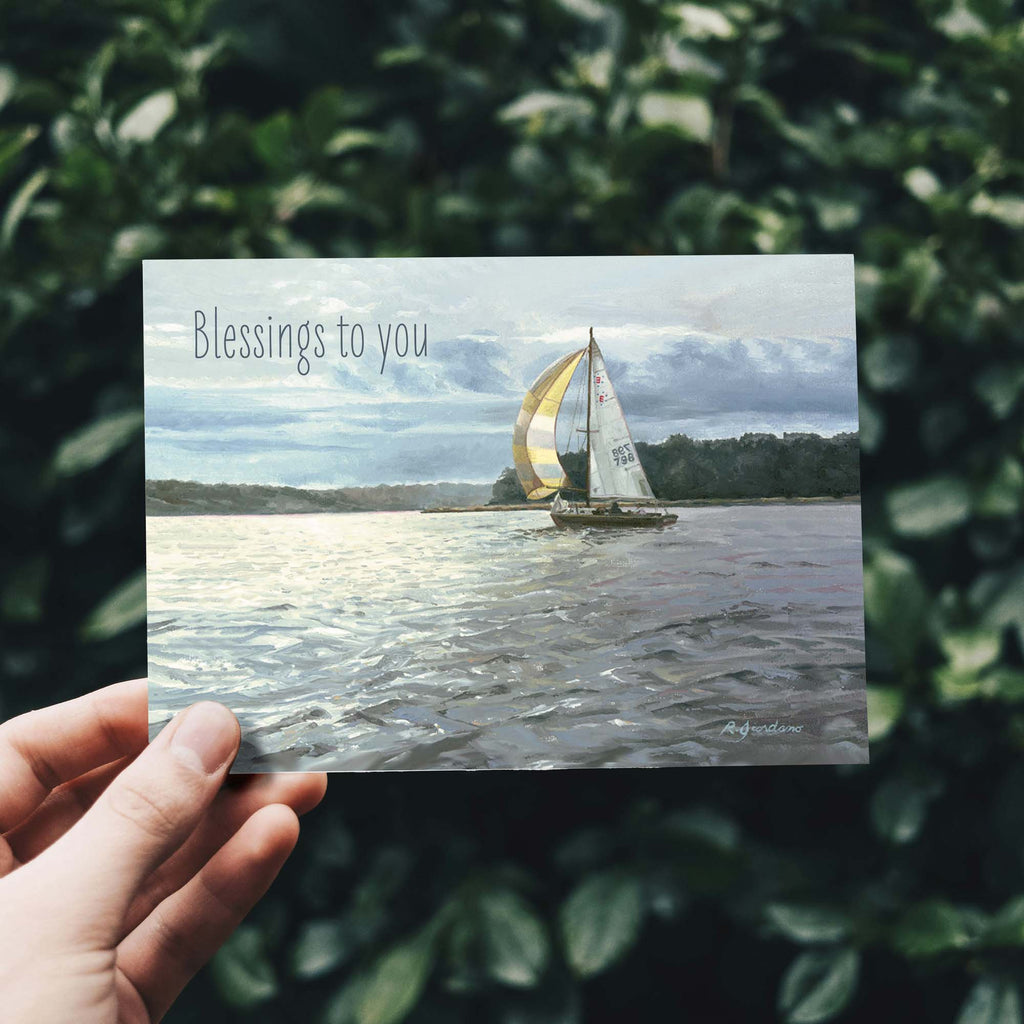 Hand holding a postcard with a sailboat painting and 'Blessings to you' text, against a leafy green background.
