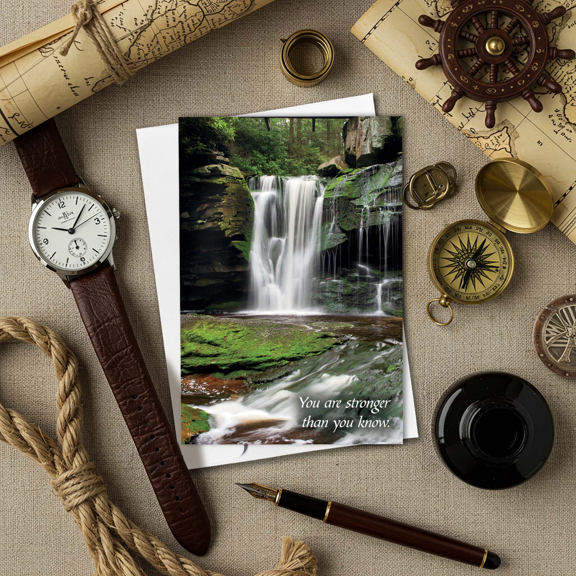 Watch, pen, and compass on a textured surface with a card featuring a waterfall and motivational quote.