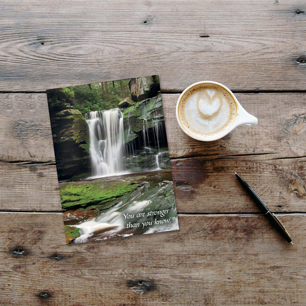 Two greeting cards with a waterfall design on a wooden surface next to a cup of coffee.