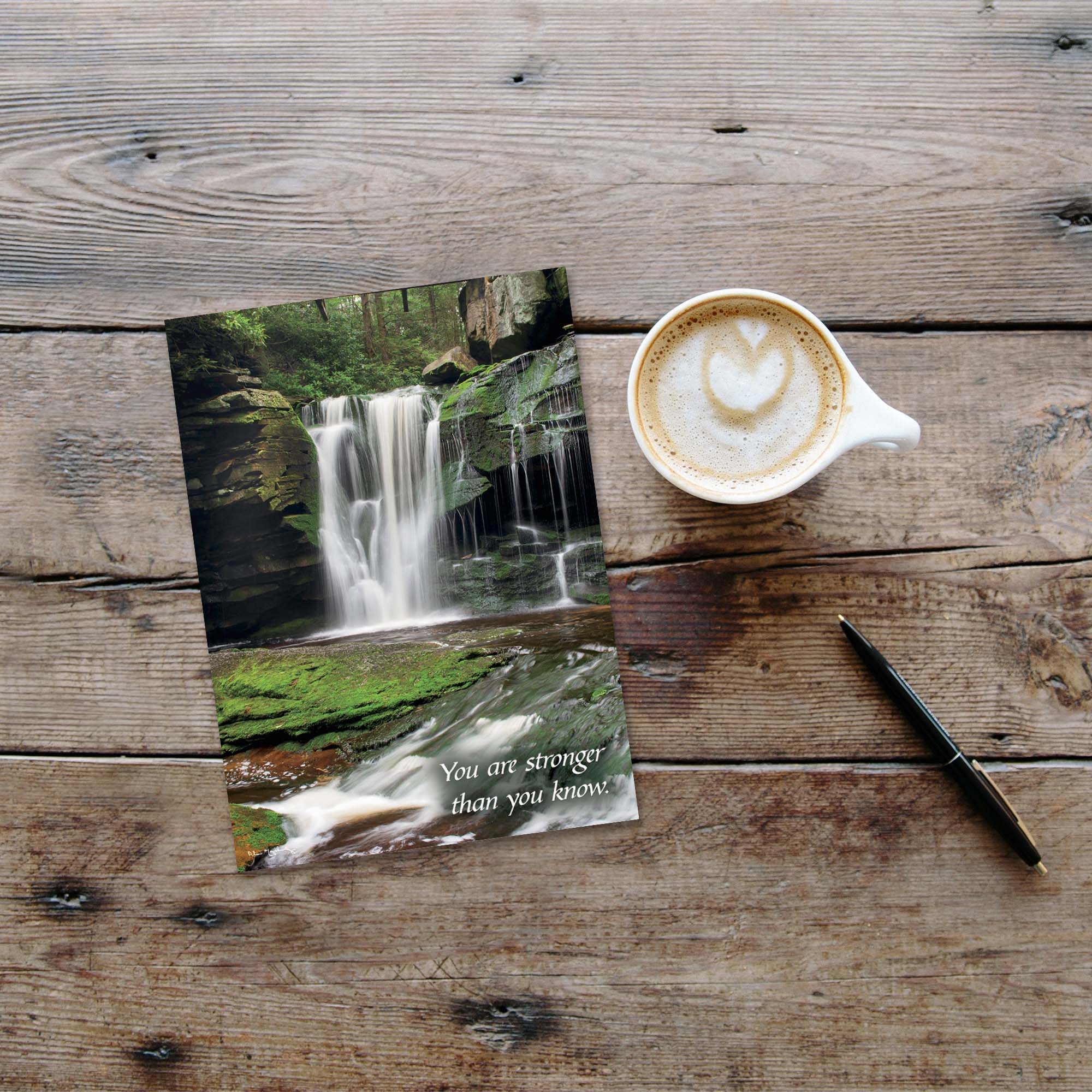 Two greeting cards with a waterfall design on a wooden surface next to a cup of coffee.