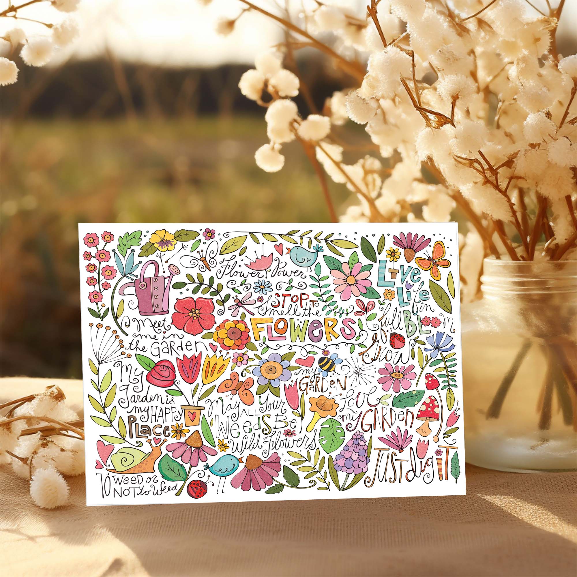 Colorful floral and garden-themed card with decorative text on a blurred natural background.