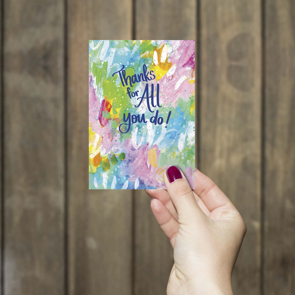Hand holding a colorful card with 'Thanks for All you do!' text against a wooden background