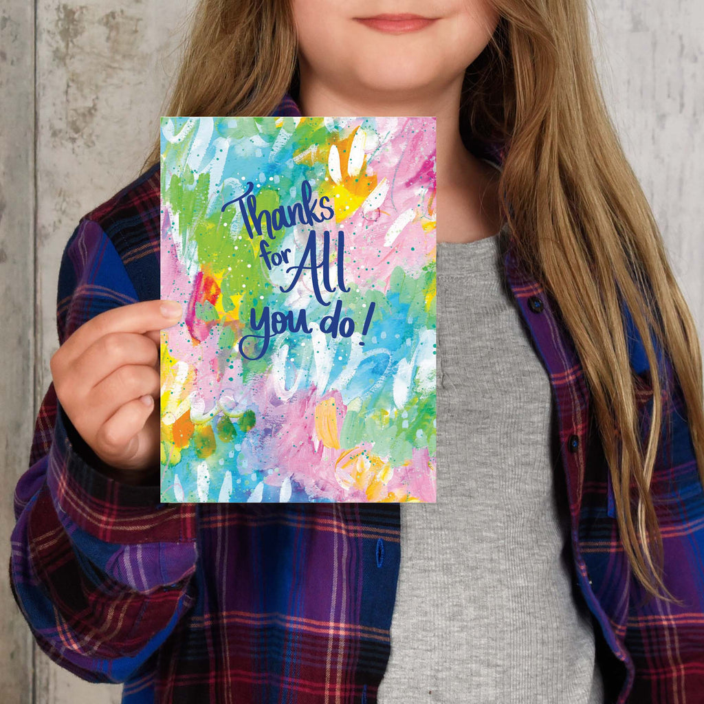 Person holding a colorful card with 'Thanks for All you do!' text against a concrete wall.