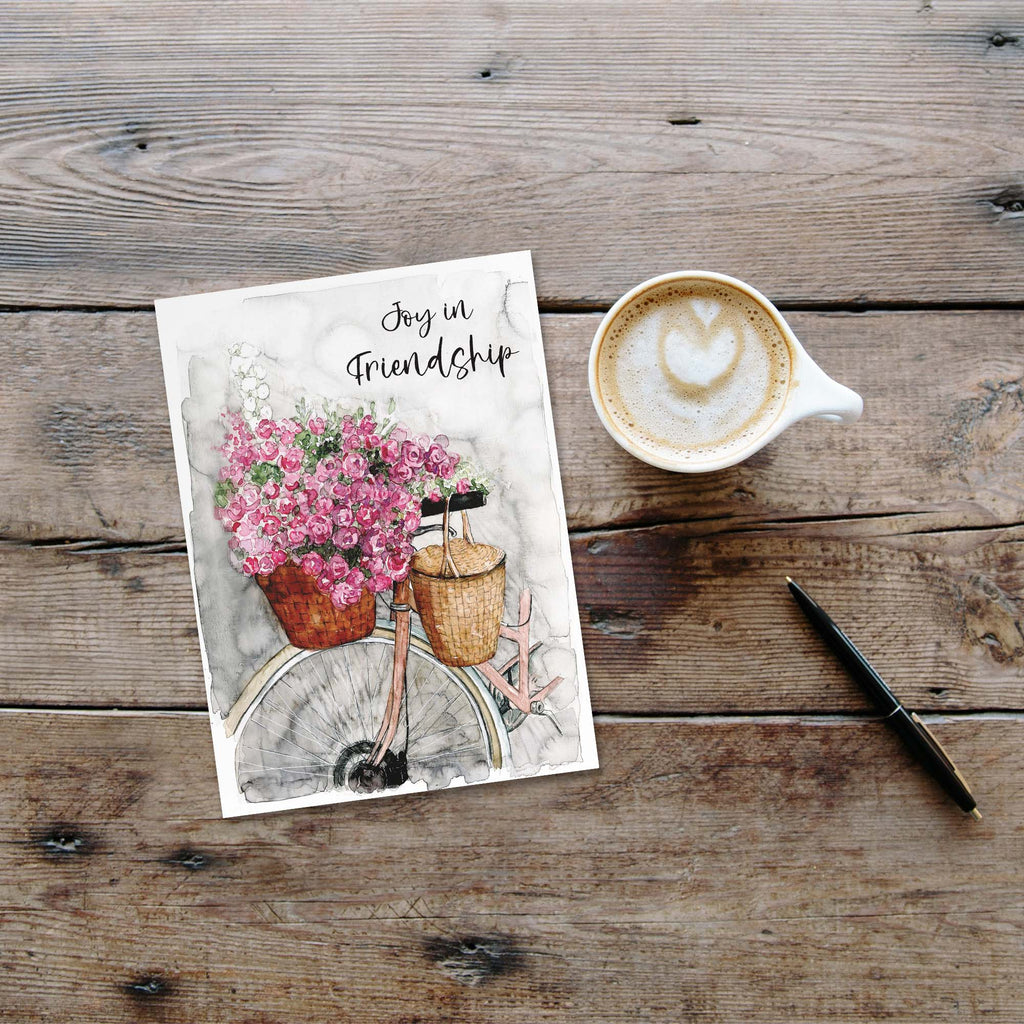 Greeting card with flowers and 'Joy in Friendship' text on a wooden surface with a coffee cup and pen.