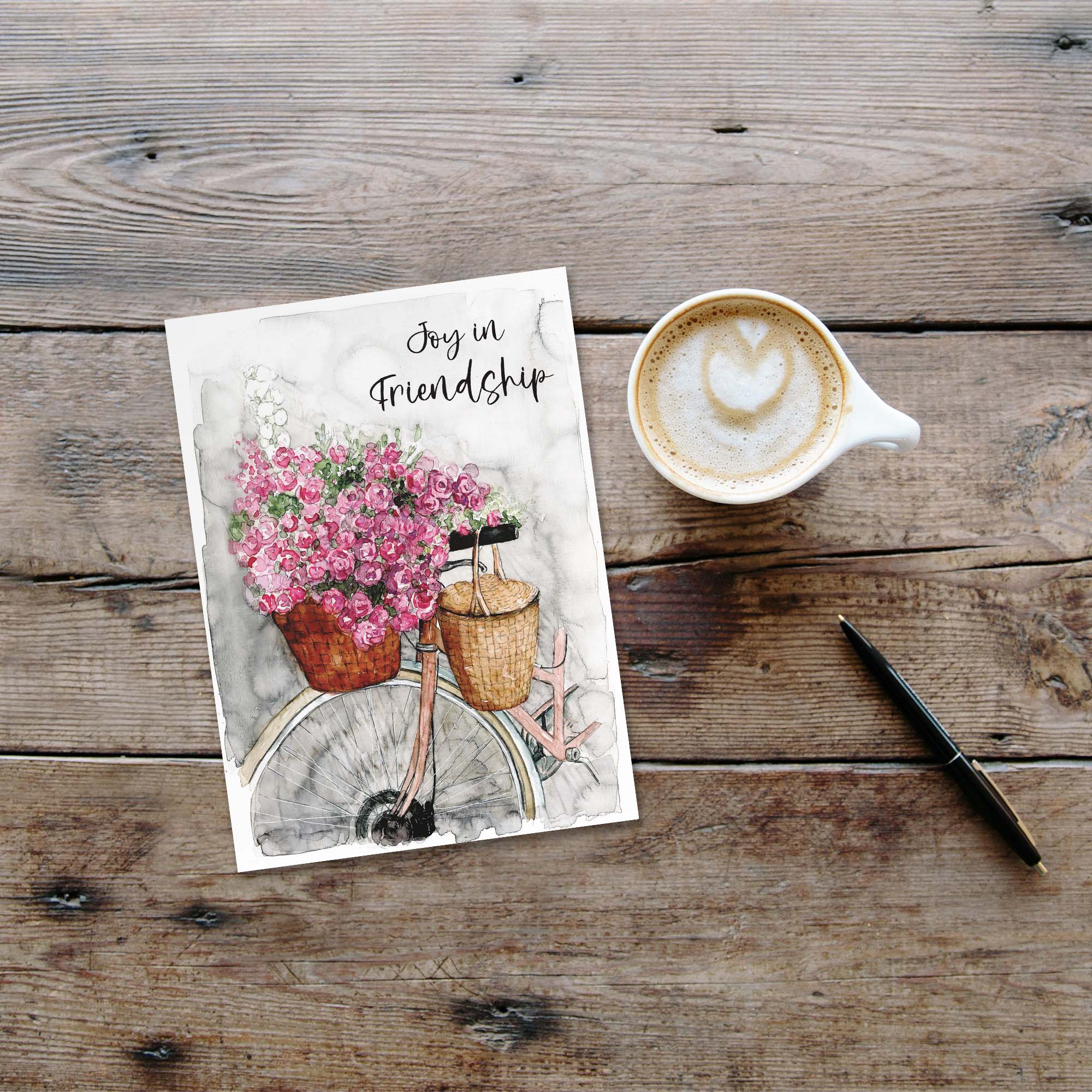 Greeting card with flowers and 'Joy in Friendship' text on a wooden surface with a coffee cup and pen.