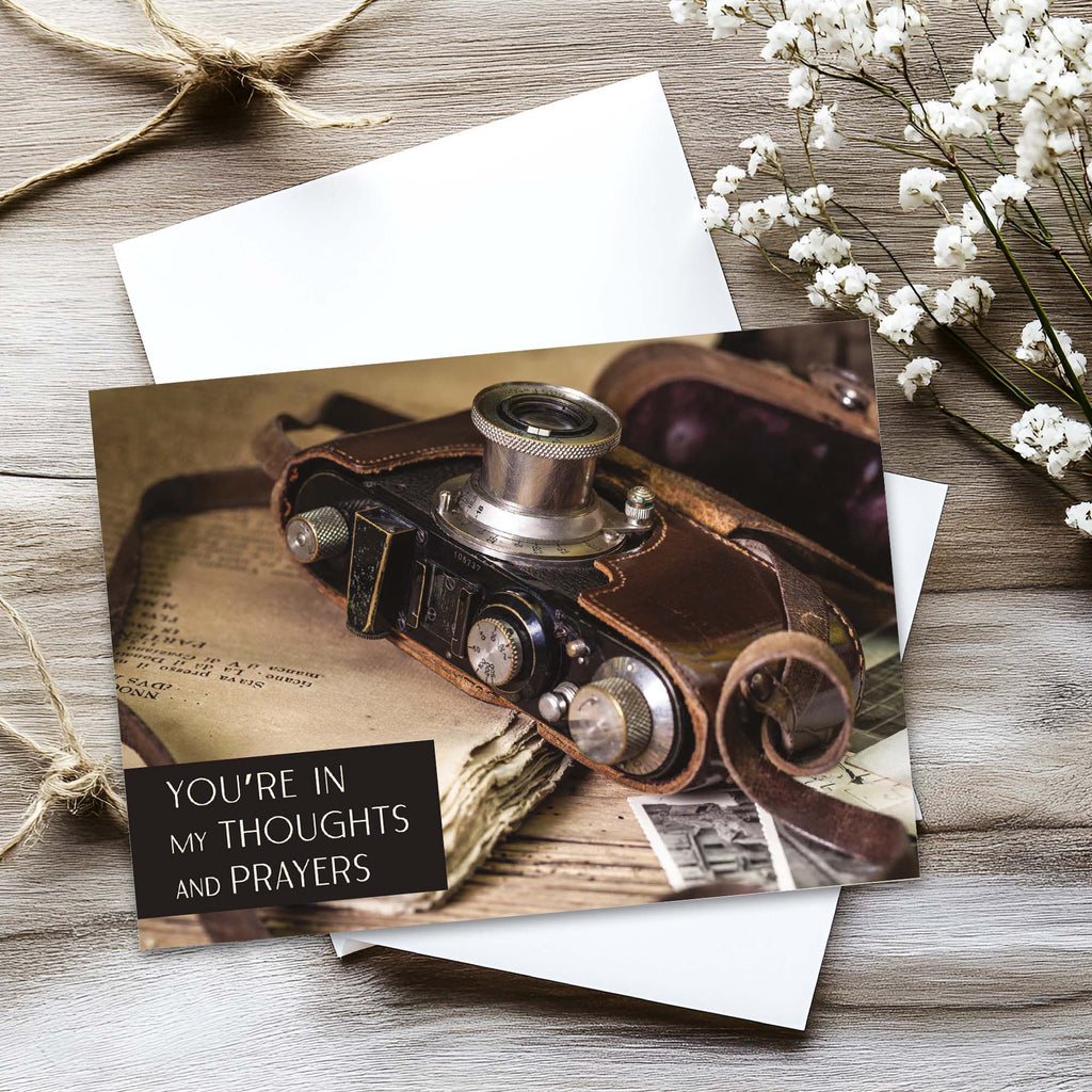 Vintage camera-themed card with 'You're in my thoughts and prayers' text on a wooden surface with flowers.