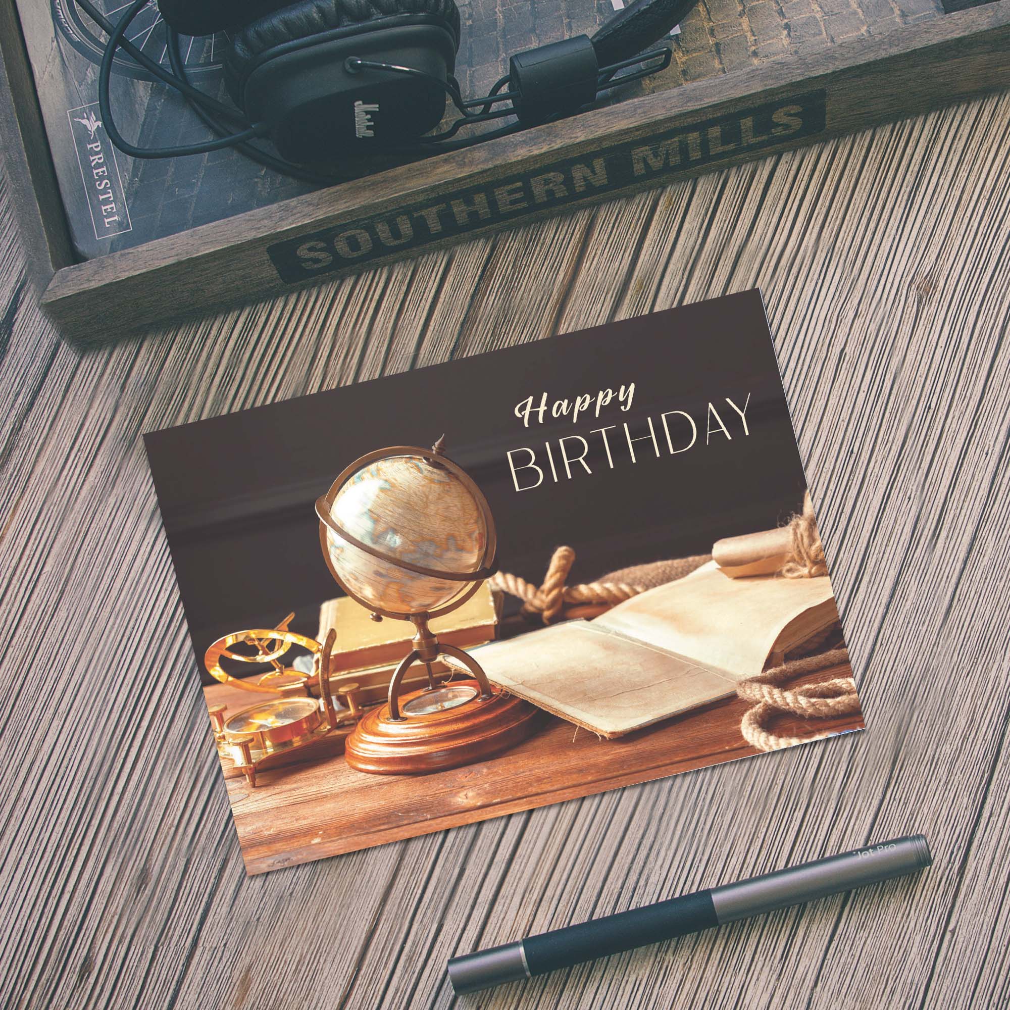 Birthday card with a globe and books on a wooden surface, with a pen and framed picture in the background.