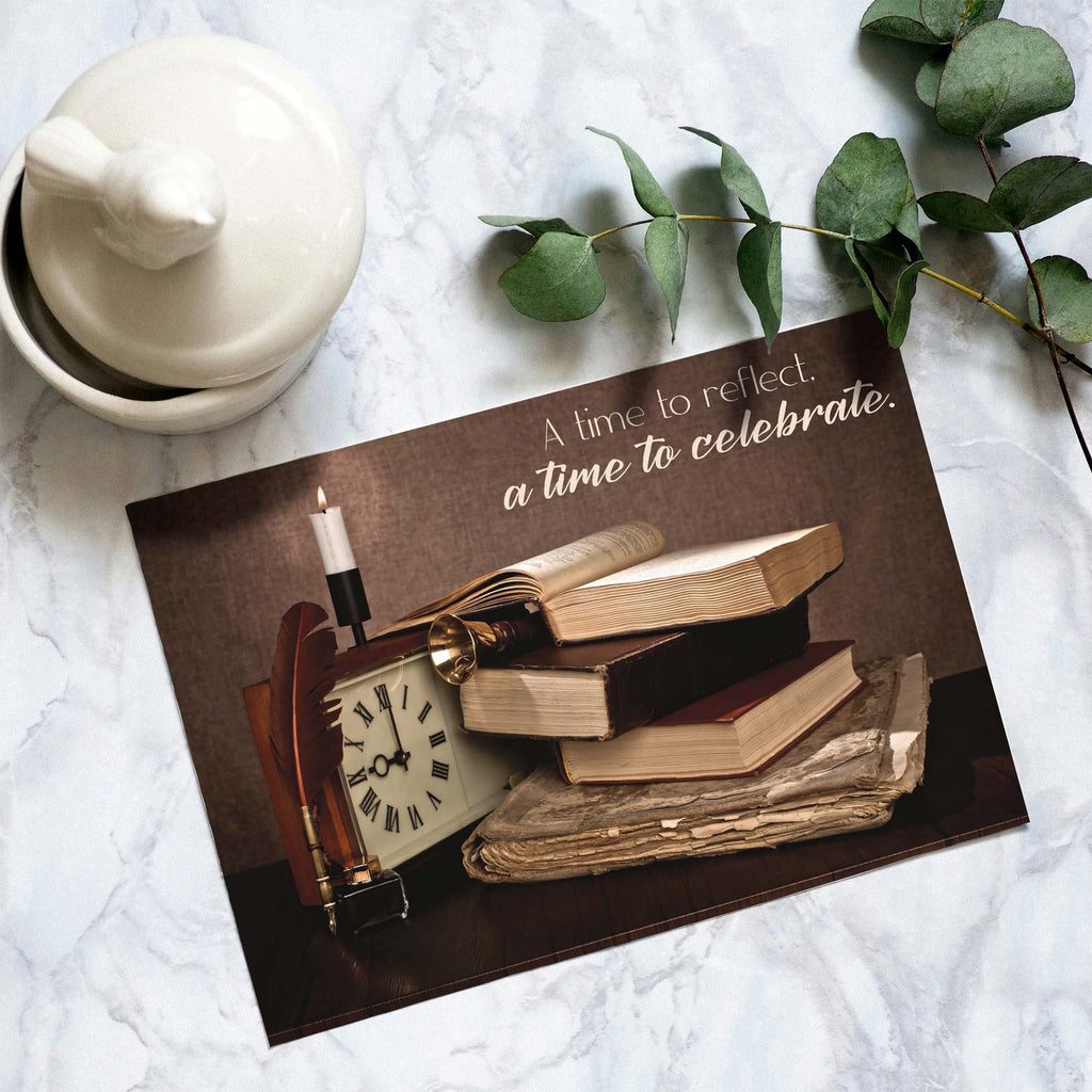 Decorative card with books, clock, and candle on a marble surface with a white teapot and green leaves.