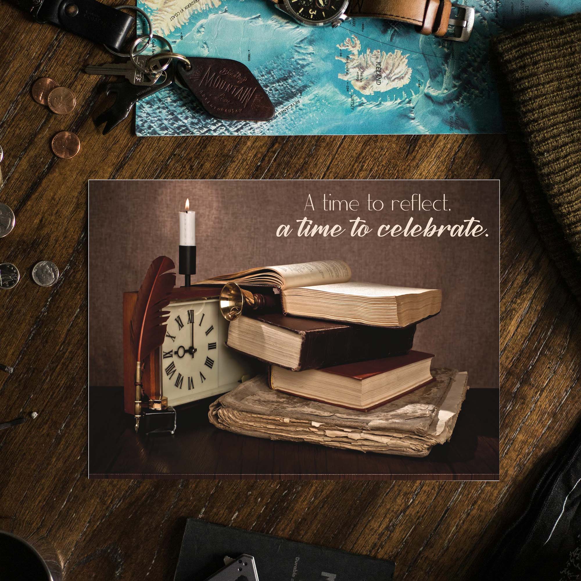 Decorative card with books, clock, and candle on a wooden surface with text 'A time to reflect, a time to celebrate'.