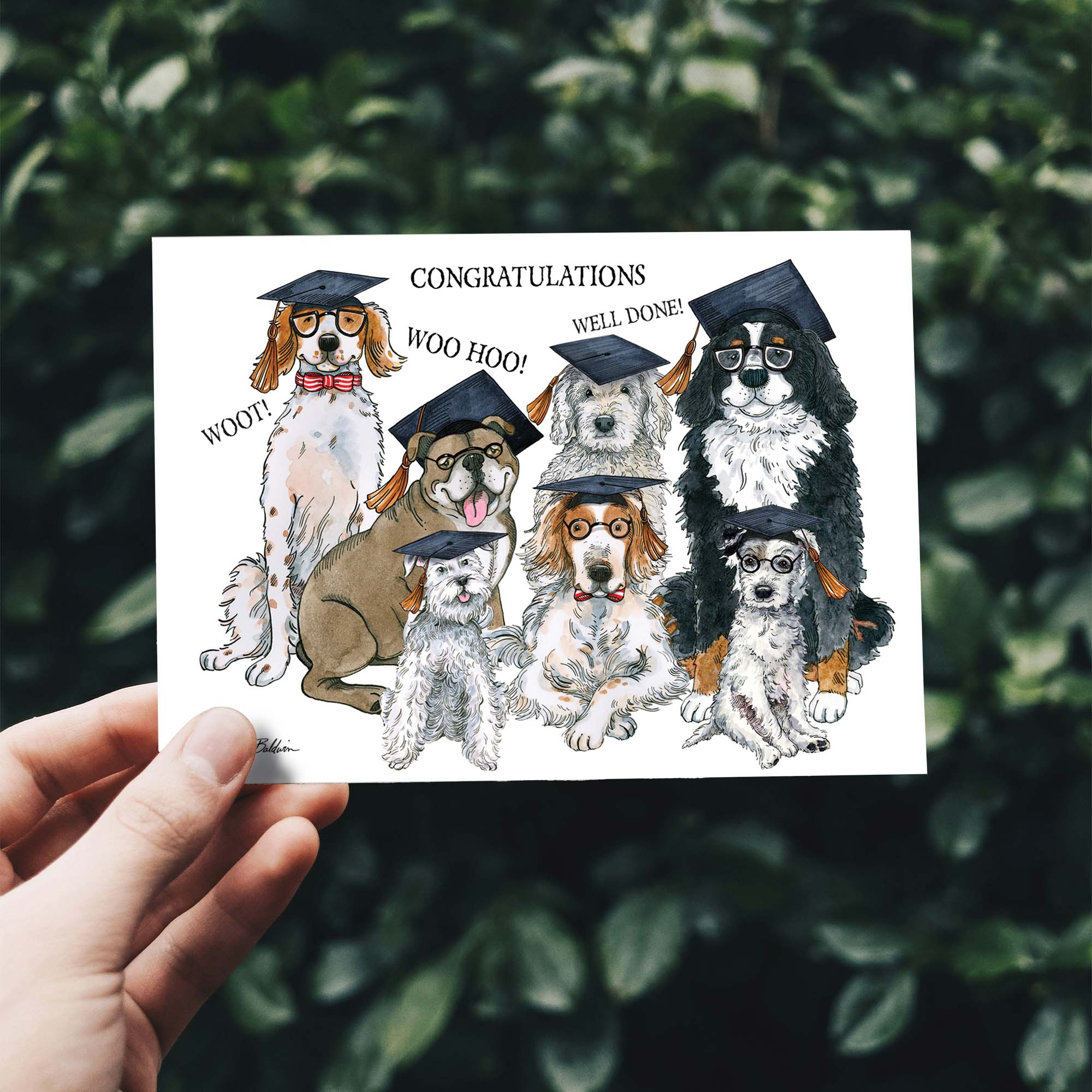 Hand holding a card with cartoon dogs in graduation caps and tassels against a green leafy background.