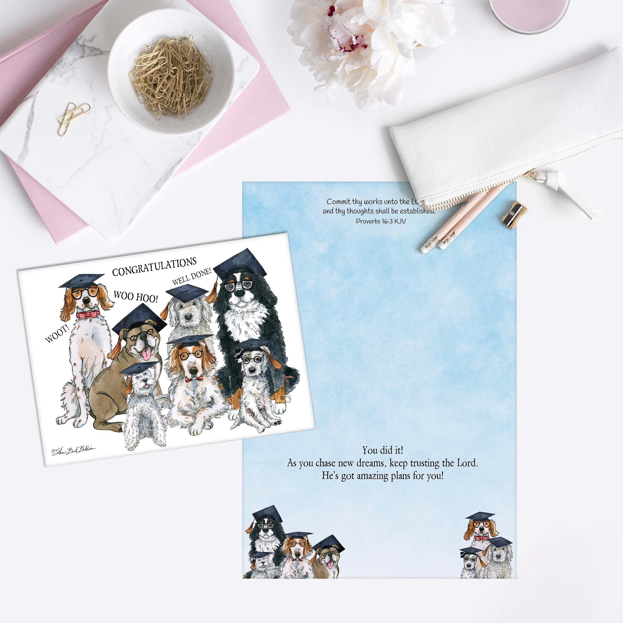 Greeting card with illustrations of dogs in graduation caps and gowns on a white surface with stationery items.