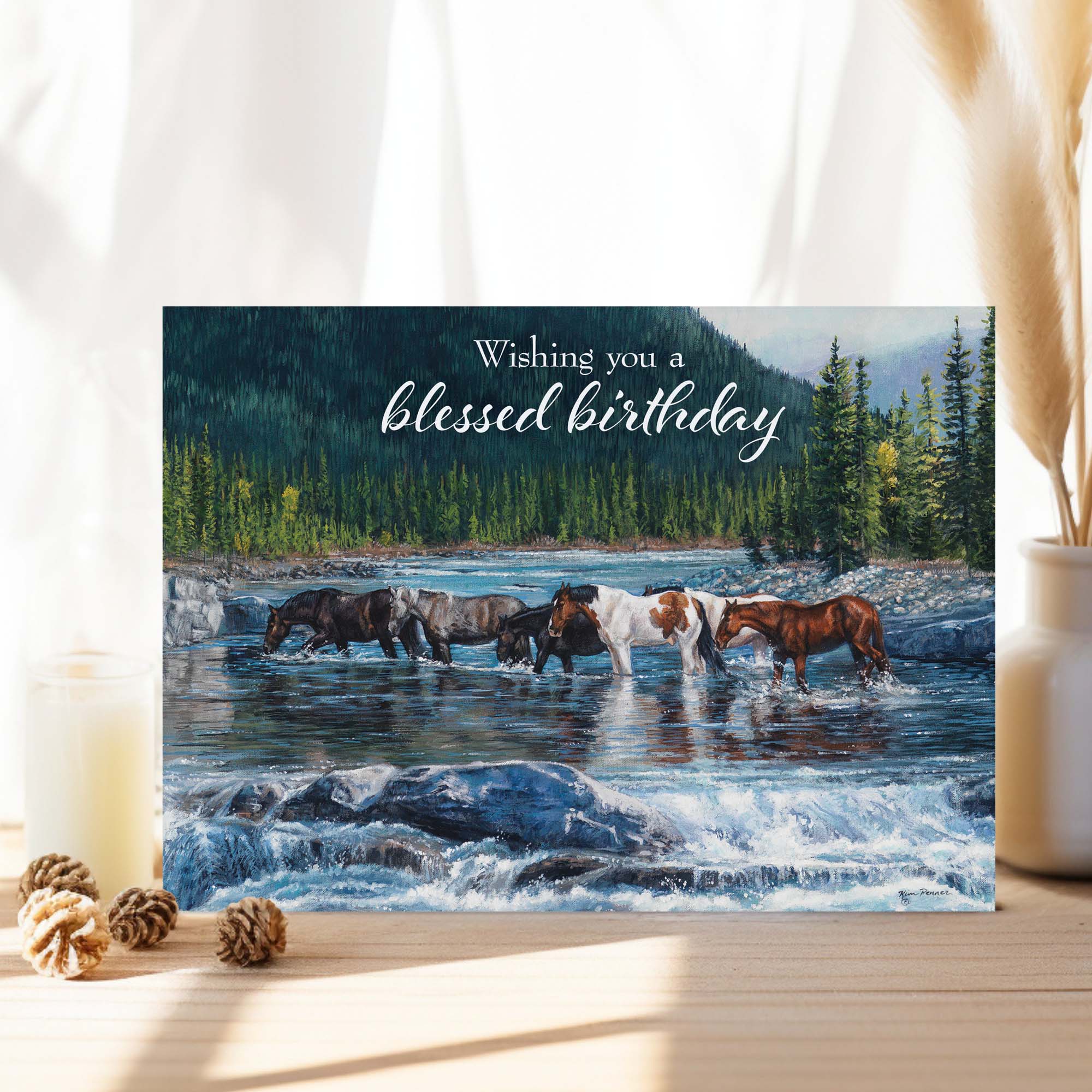 Greeting card with horses crossing a river and 'Wishing you a blessed birthday' text, placed on a surface with decorative items.