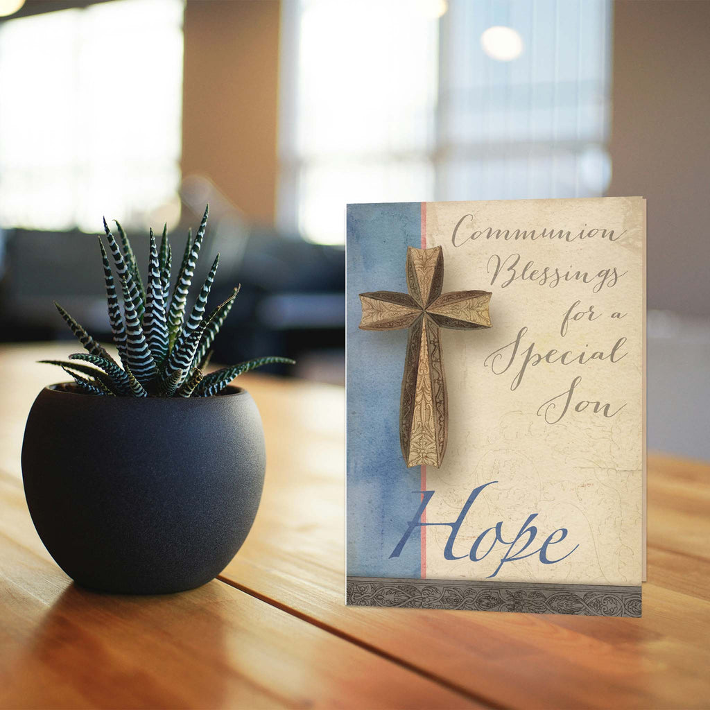 Decorative card with cross design and text on a wooden surface next to a potted plant.