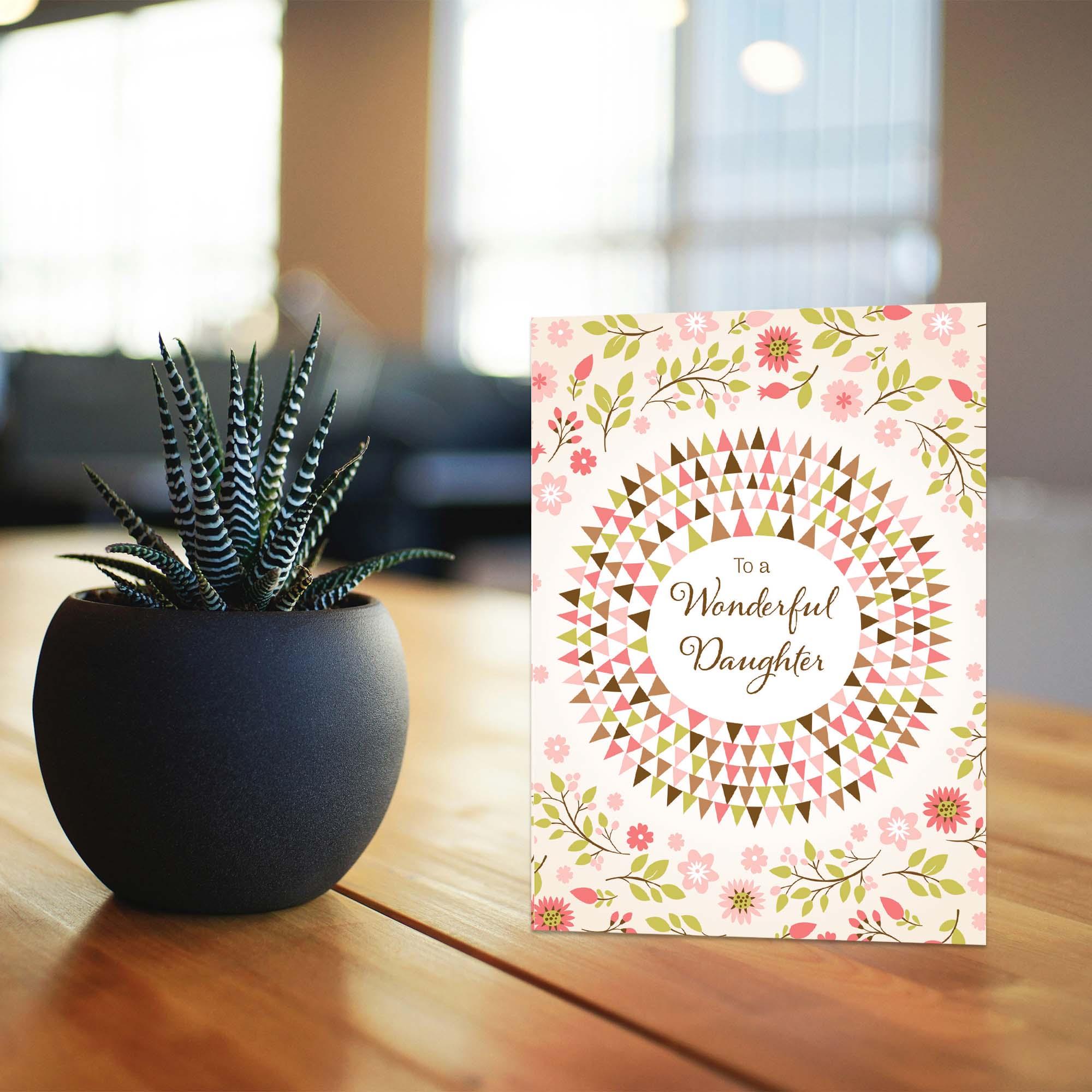 Greeting card with floral design and text 'To a Wonderful Daughter' next to a potted plant on a wooden surface.
