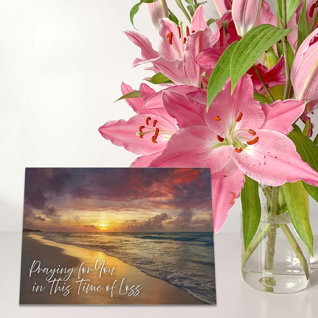 Pink lilies in a clear vase with a card featuring a sunset beach scene.