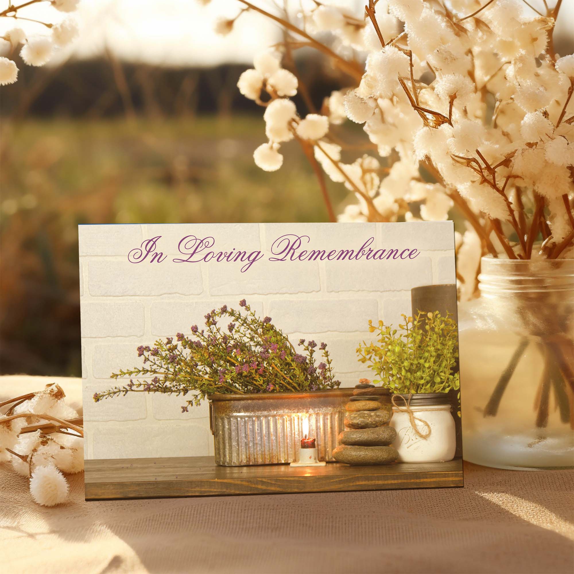 Memorial card with floral arrangement, candle, and stones on a wooden stand with cotton plants in the background.