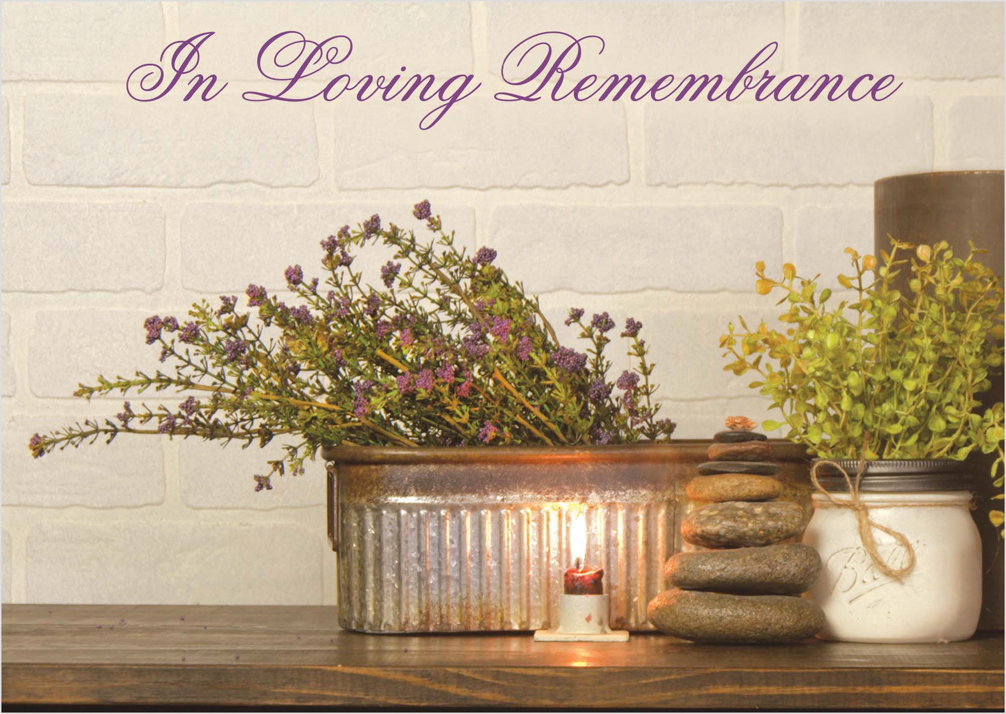 Memorial arrangement with flowers, candles, and stones on a wooden surface against a light stone wall.