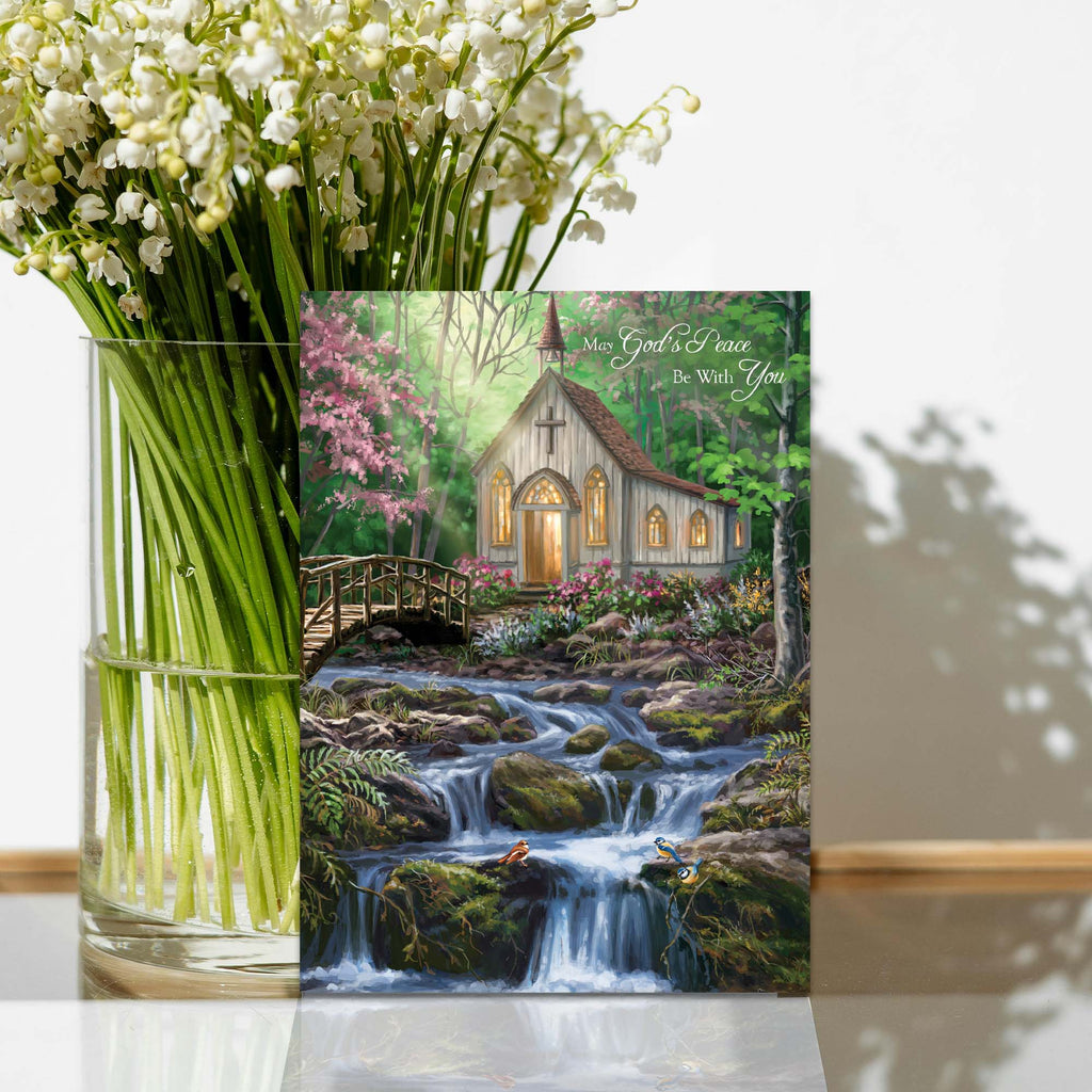 Vase of flowers with a scenic greeting card featuring a church by a stream.