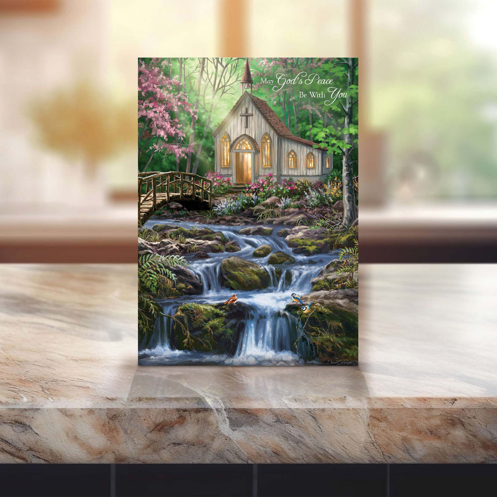 Framed artwork of a small church by a stream with pink flowers on a marble surface.