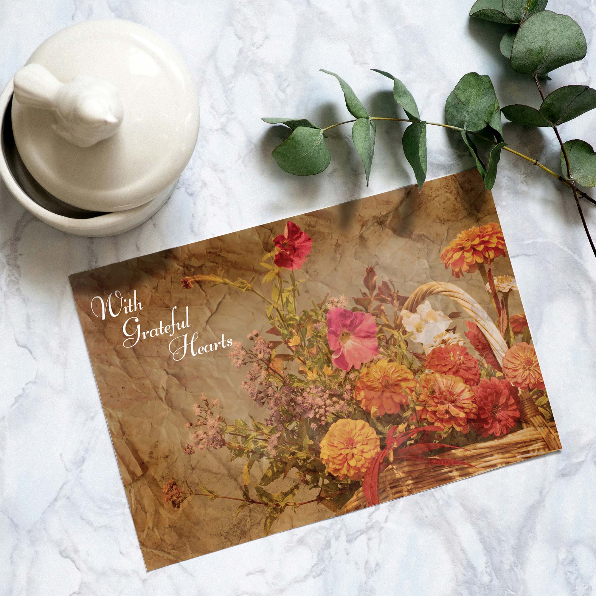Floral card with 'With Grateful Hearts' text on a marble surface with a teapot and eucalyptus leaves.