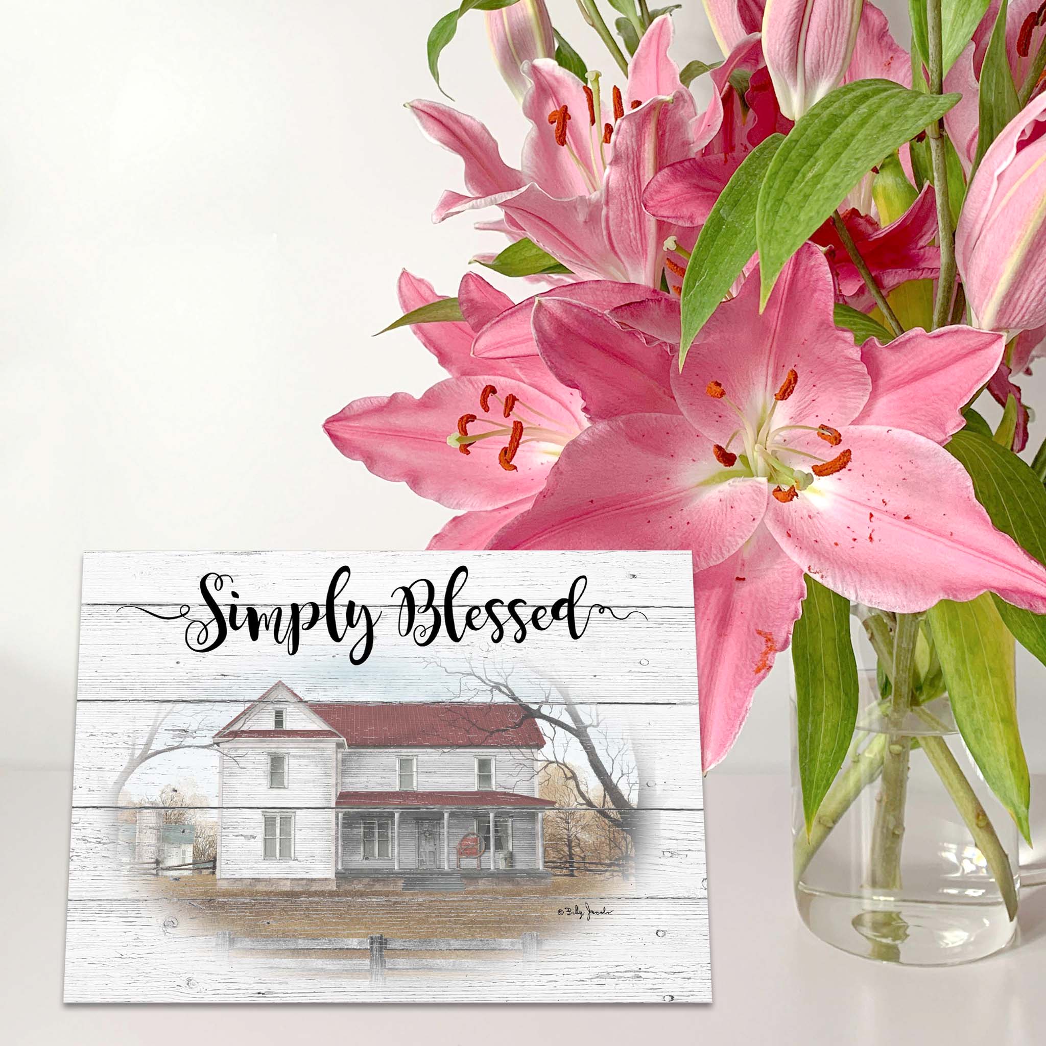 Pink lilies in a vase with a sign reading 'Simply Blessed' featuring a house illustration.