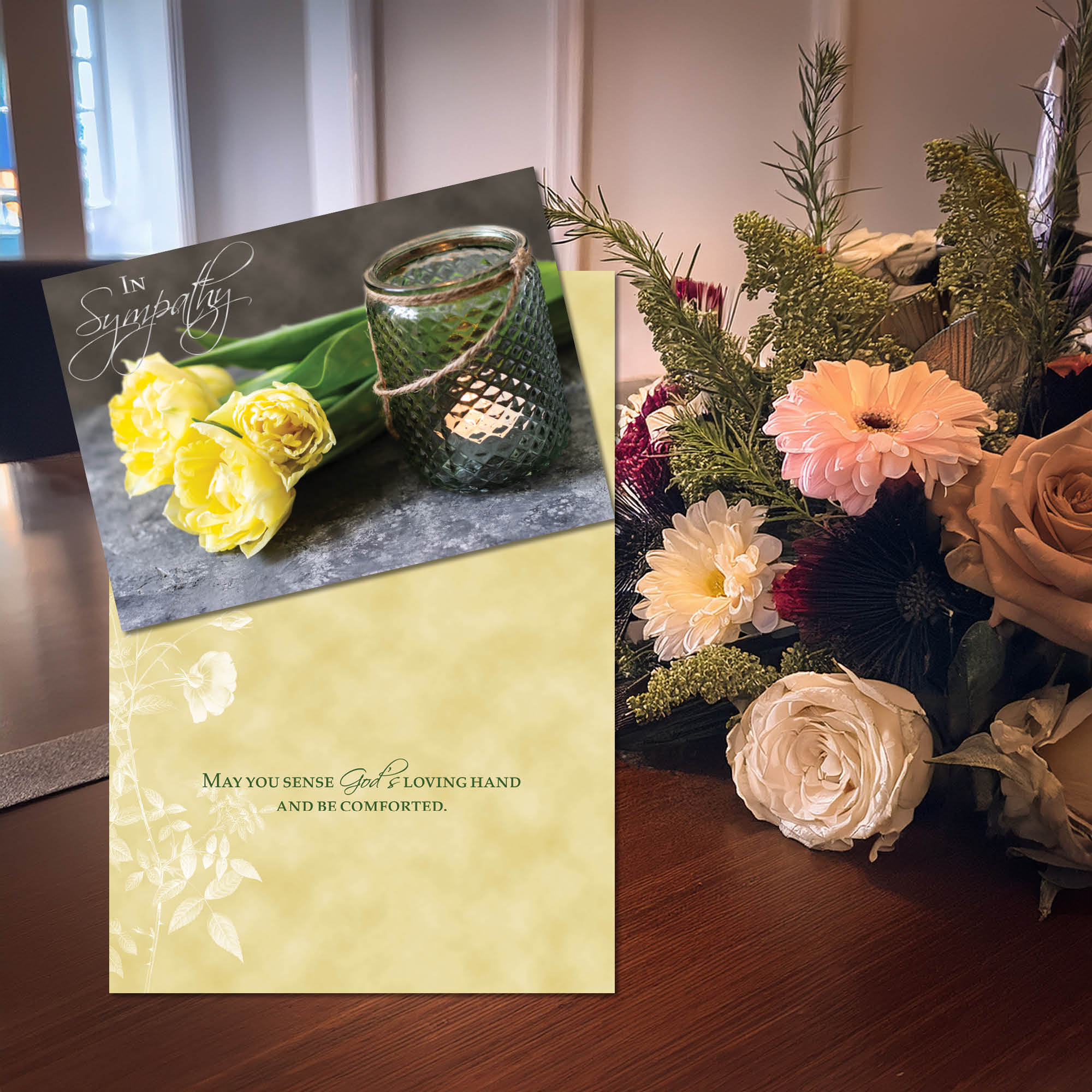 Sympathy card with yellow roses and a candle, next to a bouquet of flowers on a wooden surface.